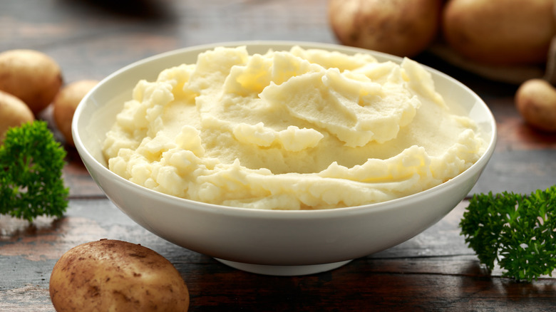 Mashed potatoes in a white bowl surrounded by raw potatoes