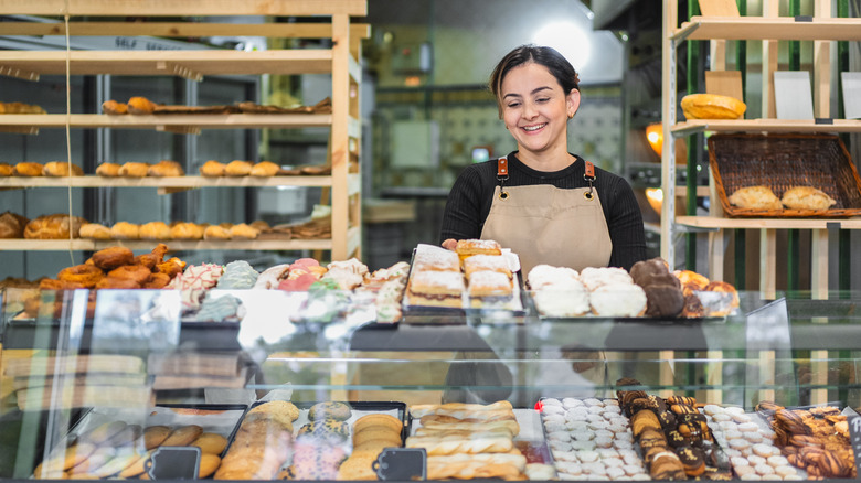A young female baker arranges fresh pastries in a display case at a bakery.