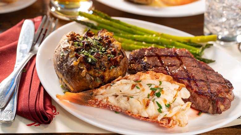 White dinner plate with steak, lobster, loaded baked potato and asparagus on table