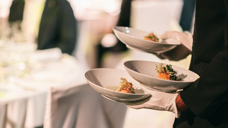 A waiter with white gloves carrying three plates of food in a Michelin starred restaurant