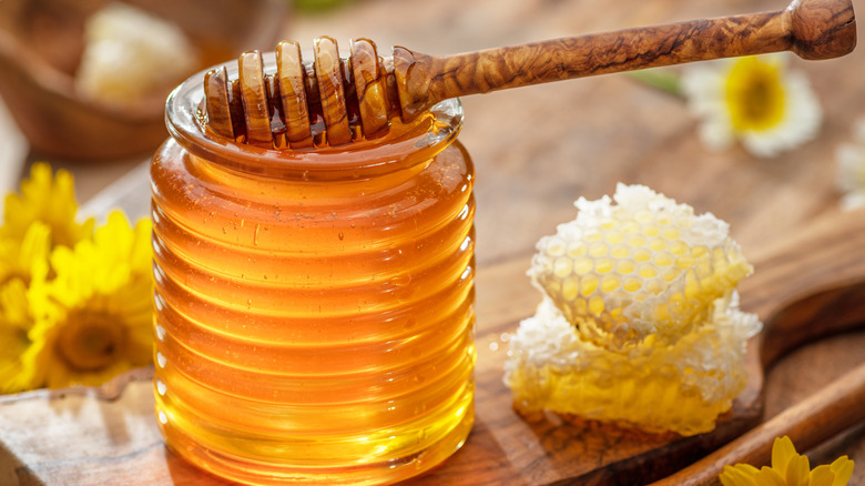 Honey in a glass jar with a dipper, honeycomb, and flowers