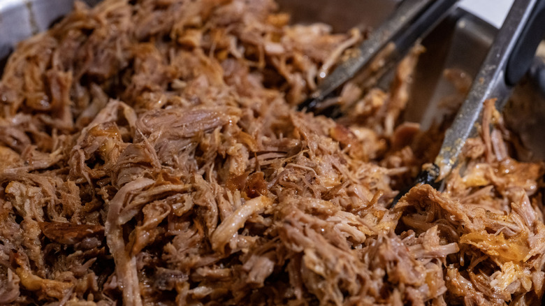 A pan of pulled pork with metal tongs