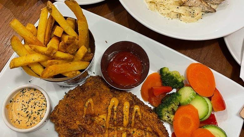 A plate of food, with french fries, fried fish and mixed vegetables, next to two ramekins of ketchup and sauce