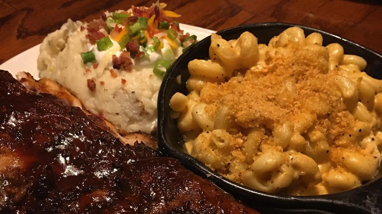 A plate full of ribs, mac and cheese and mashed potatoes topped with bacon, onions, and cheese