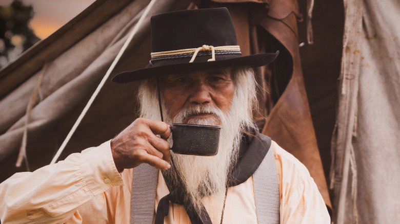 An old rustic man drinking a cup of coffee at a campsite
