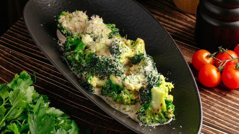 An oblong dark bowl with cheesy cauliflower in it with herbs on the left and tomatoes on the right