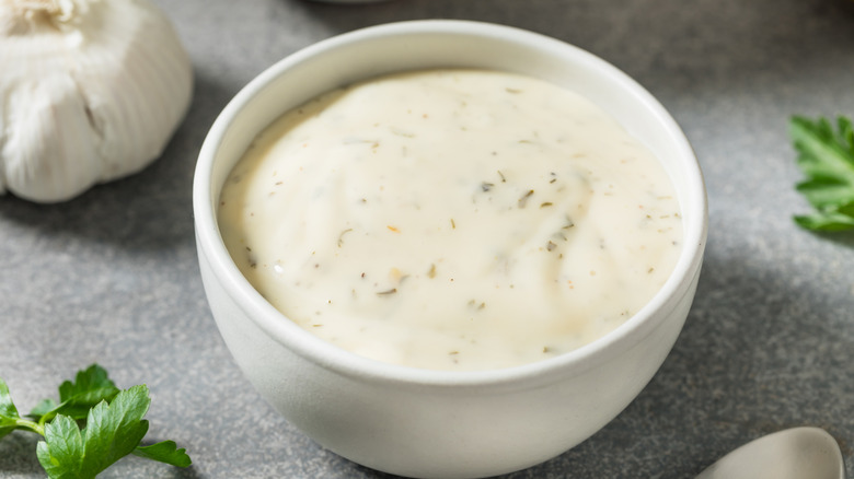 A bowl of ranch dressing in a white ramekin on a gray surface surrounded by a head of garlic and parsley sprigs