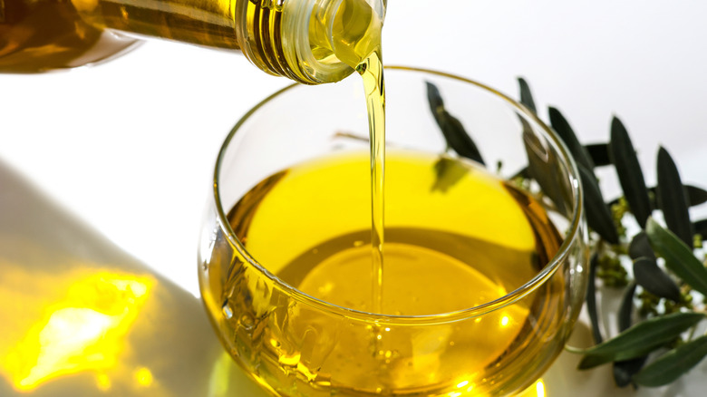 Olive oil pouring from a bottle into a round glass bowl on a white background with olive leaves on the right