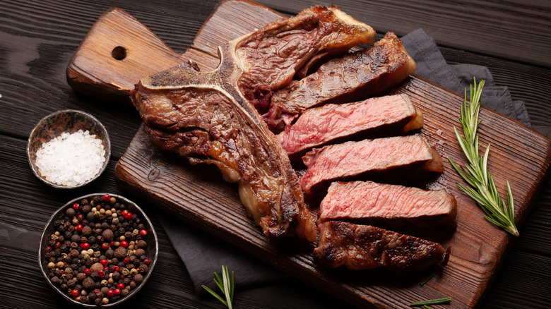 A sliced T-bone steak on a wood cutting board with a bowl of salt, a bowl of peppercorns, and a sprig of rosemary
