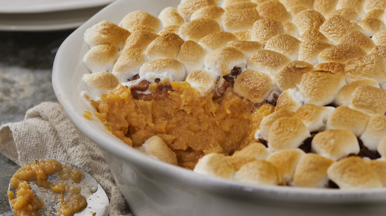 A close-up of sweet potato casserole with marshmallows on top in a round dish with a napkin and spoon on the left