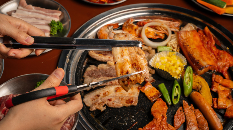 Hands using scissors to cut grilled pork on a Korean BBQ pan with corn and green peppers
