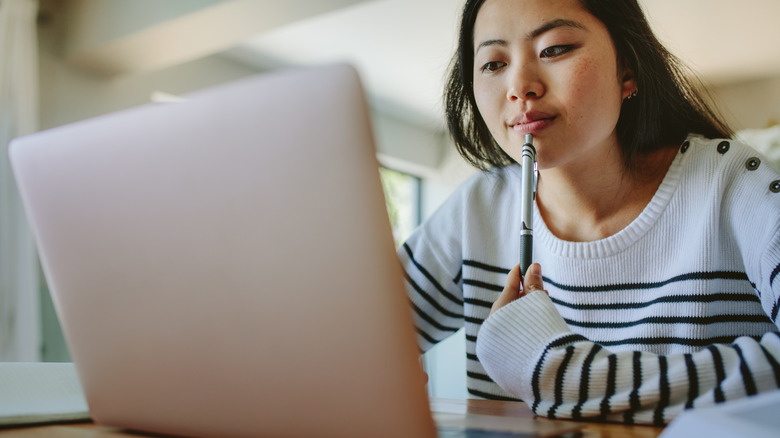 A young woman using a laptop and holding a pen with a pensive expression