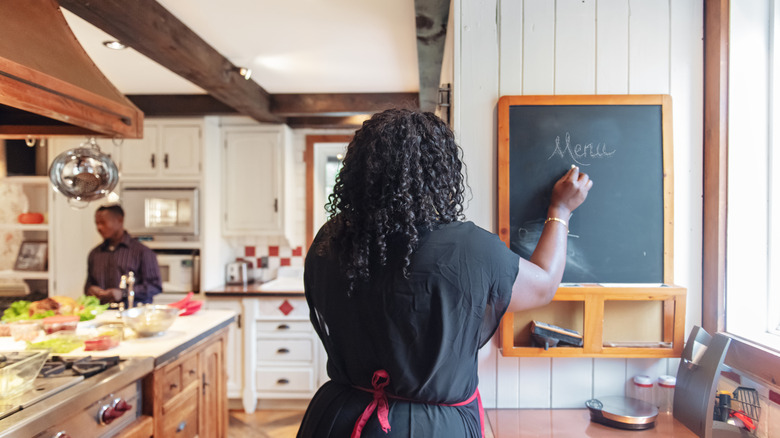 A woman photographed from behind while writing "menu" on a kitchen chalkboard