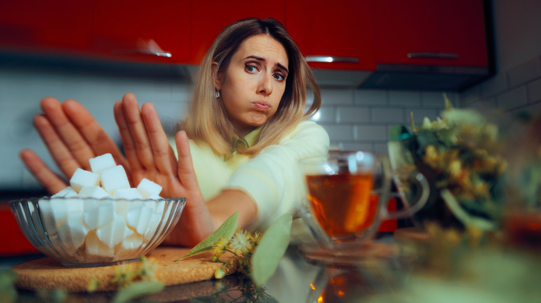 A woman in a kitchen, frowning while pushing a bowl of sugar cubes away