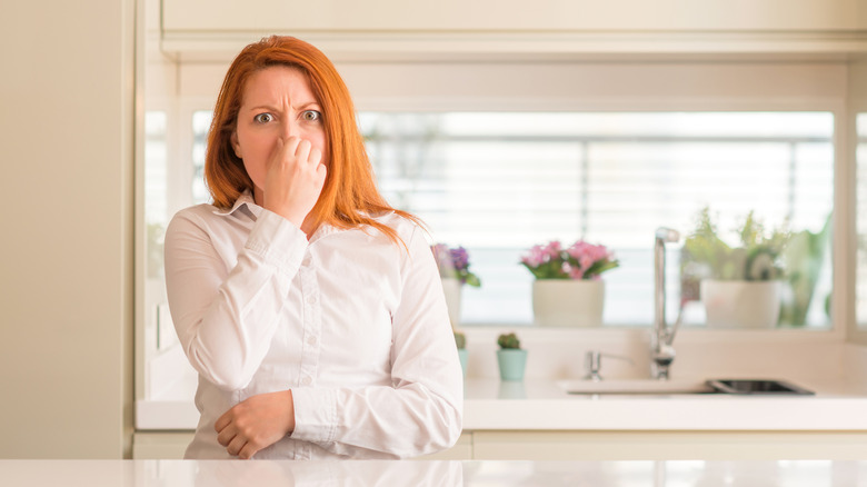 Redhead woman at kitchen smelling something stinky and disgusting, intolerable smell, holding breath with fingers on nose. Bad smells concept.