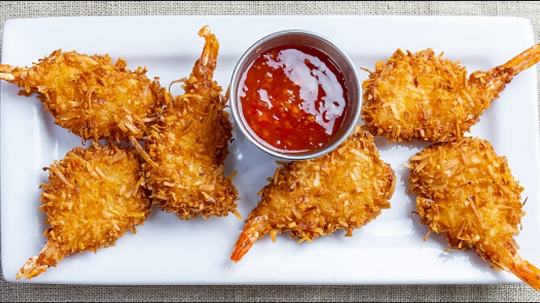 Deep fried coconut shrimp with cup of dipping sauce on a rectangular white plate