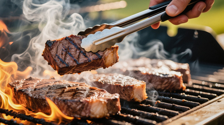 Close up of multiple steaks on grill with flames as man holds a cut of steak with tongs
