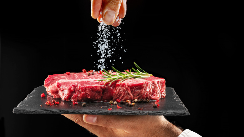 Close up of man holding black tray of steak and salting it with the other hand