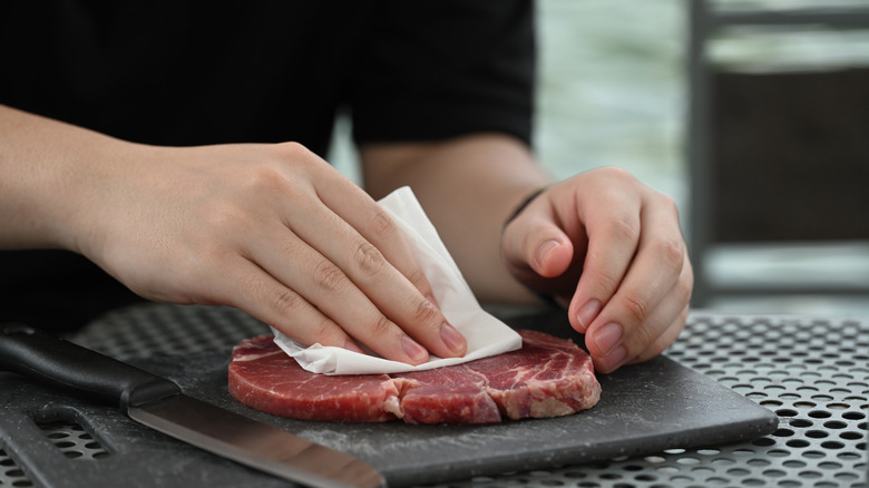 Close up of woman patting steak dry with white cloth on black cutting board next to knife