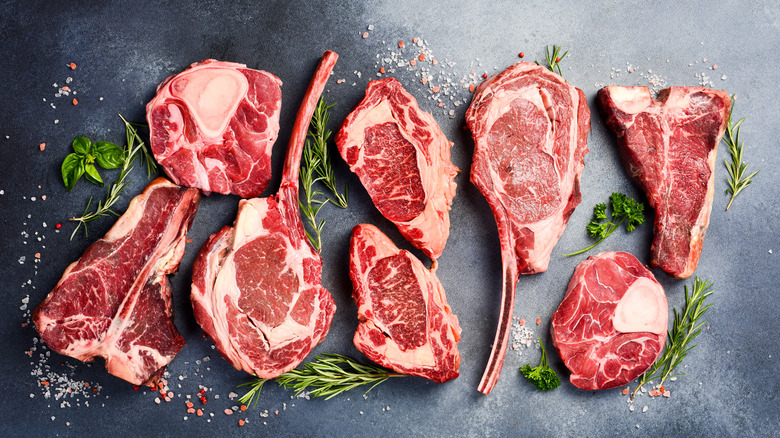Different cuts of raw beef including T-bones on black surface with herbs and salt