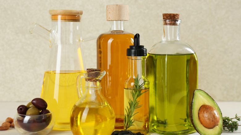 Different types of oil in various bottles next to olives, almonds, avocado, and herbs