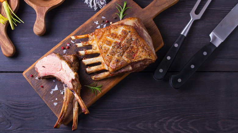 A roasted rack of lab on a board with some cutlets sliced off, next to a carving knife