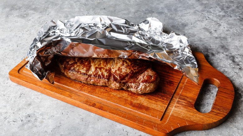 Grilled steak resting on wooden board covered with aluminum foil