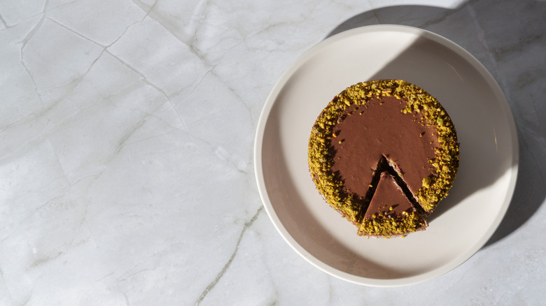 Top view of Dubai chocolate cheesecake topped with chocolate ganache and pistachios, on white plate.