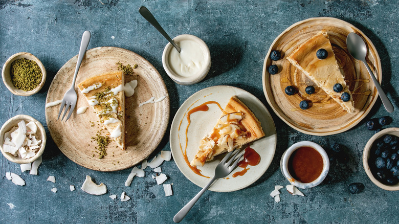Pieces of cheesecake with different toppings on ceramic plates and bowls of sauces over blue texture background.
