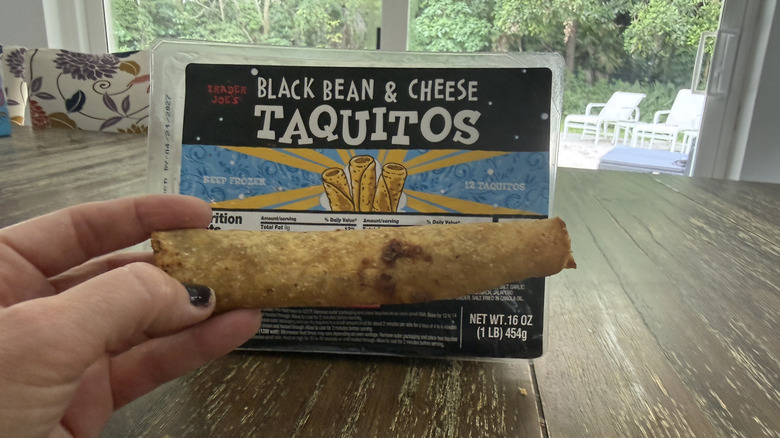 Package of Trader Joe's black bean and cheese taquitos on a wooden table and a hand holding one