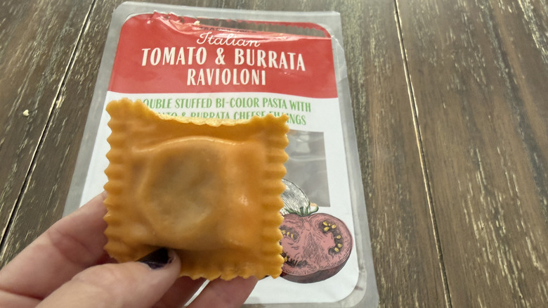 Package of Trader Joe's italian tomato and burrata raviolini on a wooden table and one held in a hand