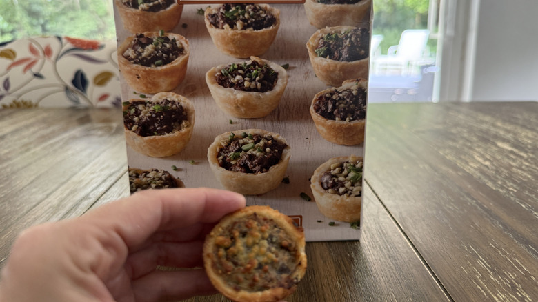Package of Trader Joe's Mushroom Tartlettes on wooden table and one held in a hand