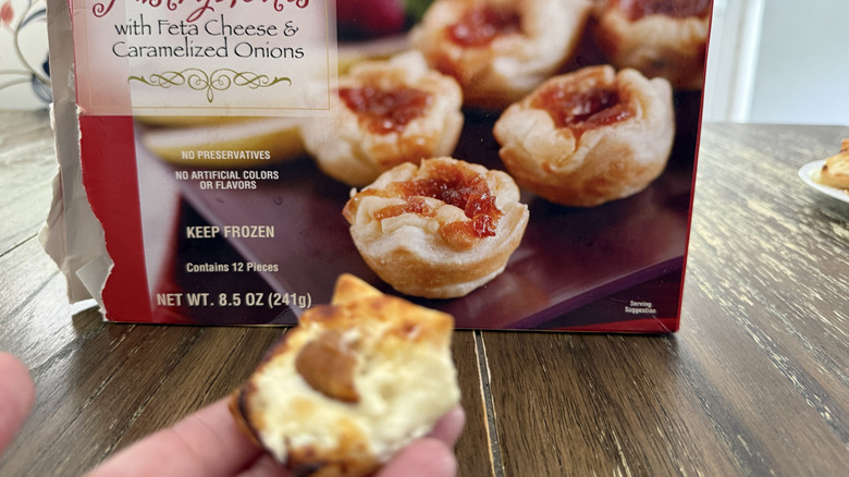 Package of Trader Joe's pastry bites with feta and caramelized onions on a wooden table and a hand holding one