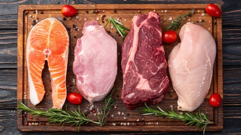 4 cuts of meat laying on a wooden cutting board surrounded by cherry tomatoes and herbs