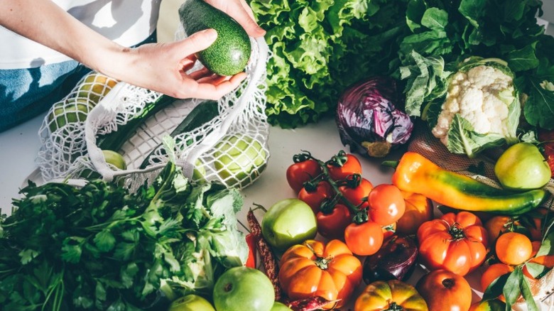 hands unpacking assorted vegetables from a mesh bag, including tomatoes, cabbage, and cauliflower