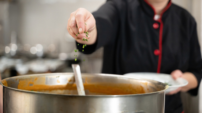 Close up of a chef in black uniform adding fresh herbs to a large pot of soup in a professional kitchen.