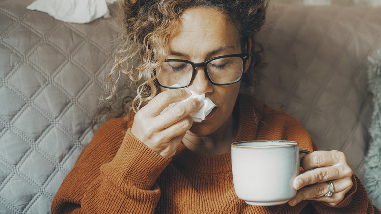 Woman looks sick and holds a tissue while drinking herbal tea medicine on sofa