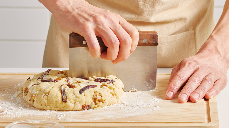 Hands using a metal bench scraper to divide raw dough on a wooden cutting board