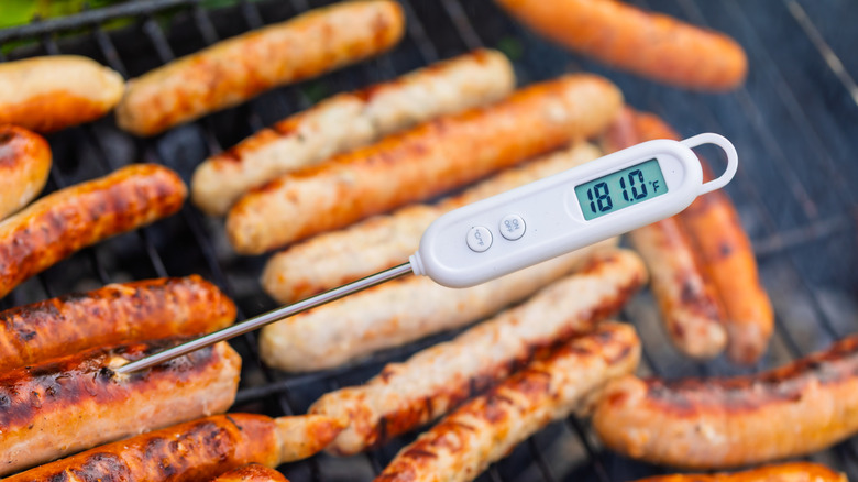 A white digital meat thermometer taking the temperature of sausages on a grill
