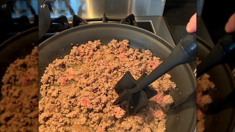 A black meat masher tool being used to grind ground beef in a hot skillet