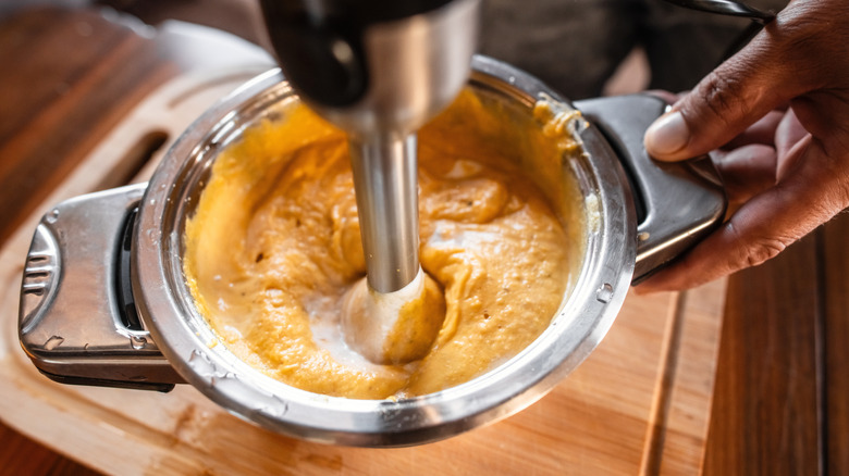 Immersion blender being used to puree food in a stainless steel pot over a wooden cutting board
