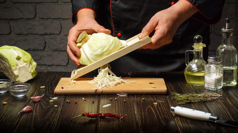 Hands shredding cabbage over a small wooden cutting board using a mandoline