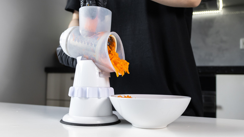 Person using a rotary grater to shred carrots into a white bowl
