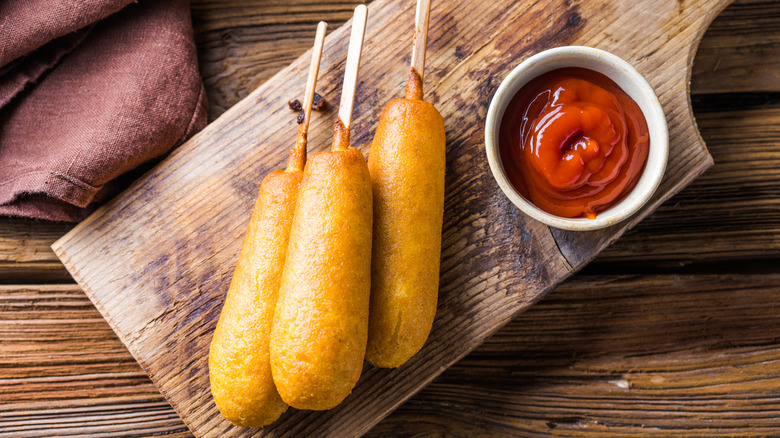Three corn dogs on a wooden board with a cup of ketchup