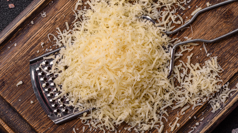 A pile of grated Parmesan cheese and a cheese grater on a dark brown wooden cutting board.