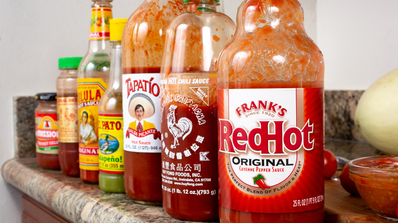 A view of a counter with a row of popular hot sauce bottles, featuring Frank's RedHot, Huy Fong Foods sriracha, Tapatio, El Pato, and Cholula.