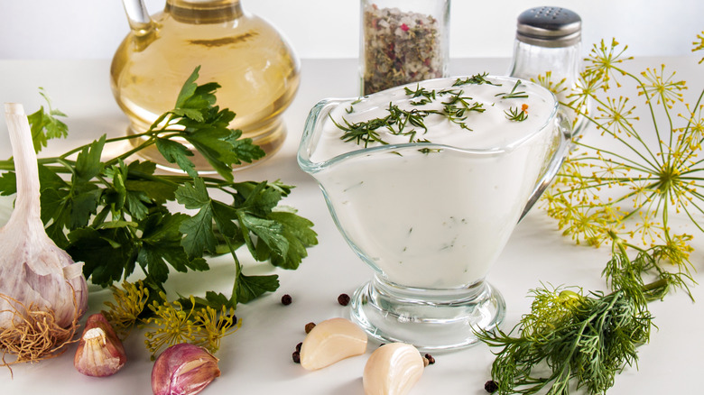 Ranch salad dressing in glass dish with garlic, herbs, parsley, dill, herbs, pepper on a white background with oil in a bottle and pepper and salt jars