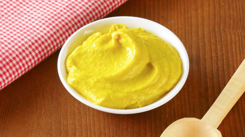 small white bowl of American yellow mustard next to red and white gingham napkin and a wooden spoon on a dark wood table.