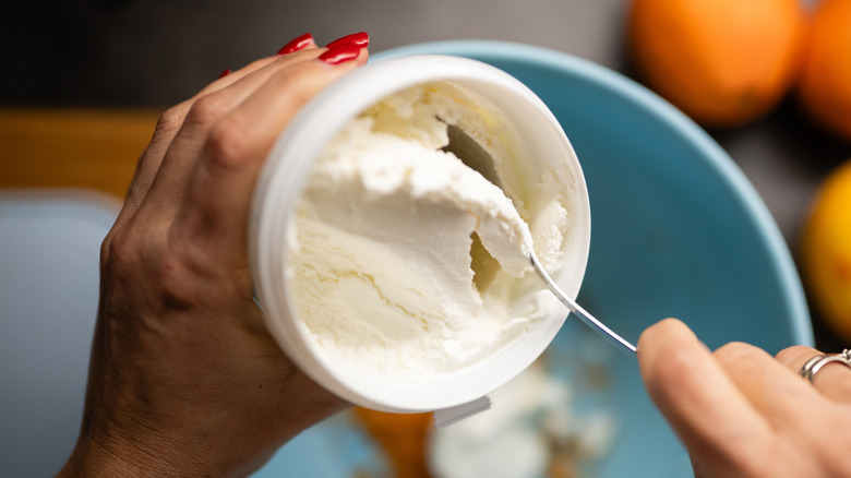 Hand scooping creamy ricotta cheese from a container into a light blue bowl
