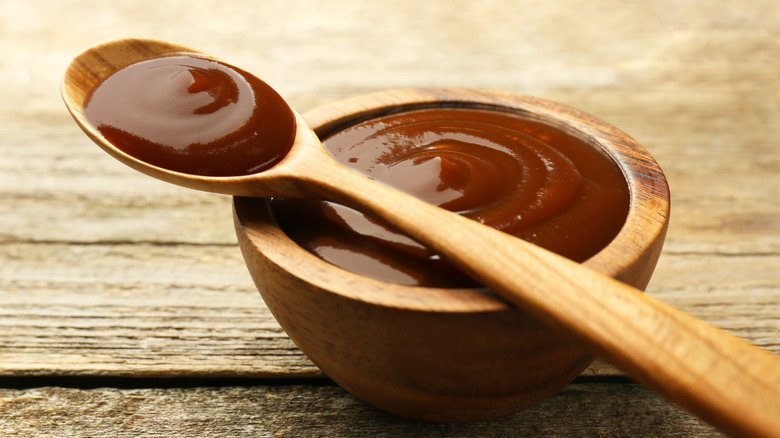 Tasty barbeque sauce in bowl and spoon on wooden table, closeup